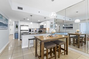 A modern kitchen with white cabinets, a central island, pendant lighting, and a dining area with a wooden table and chairs. - Grand Panama PCB 2BR Beach Views 2 Pools Sleeps 8 (Panama City Beach)