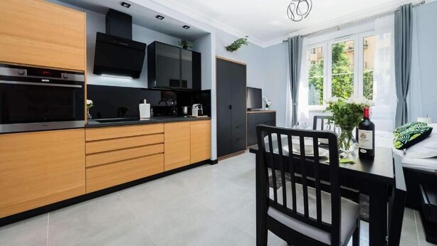 A modern kitchen with wooden cabinets, a black countertop, and a dining area with a table and chairs. - Malila Apartaments Cracow Center Dunin 3 (Kraków)