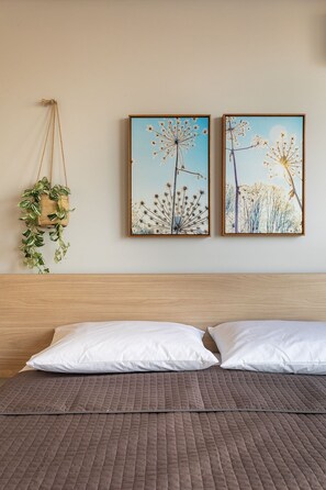 A bed with white pillows and a brown quilted bedspread, a wooden headboard, two framed pictures of flowers on the wall, and a hanging plant with green leaves.