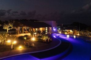 A swimming pool with a curved design, surrounded by a covered seating area and a building with a flat roof.