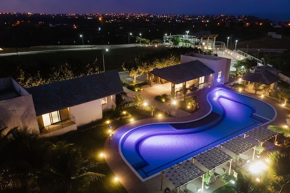 A swimming pool with a curved design, surrounded by lounge chairs and umbrellas, and adjacent to a building with a balcony.