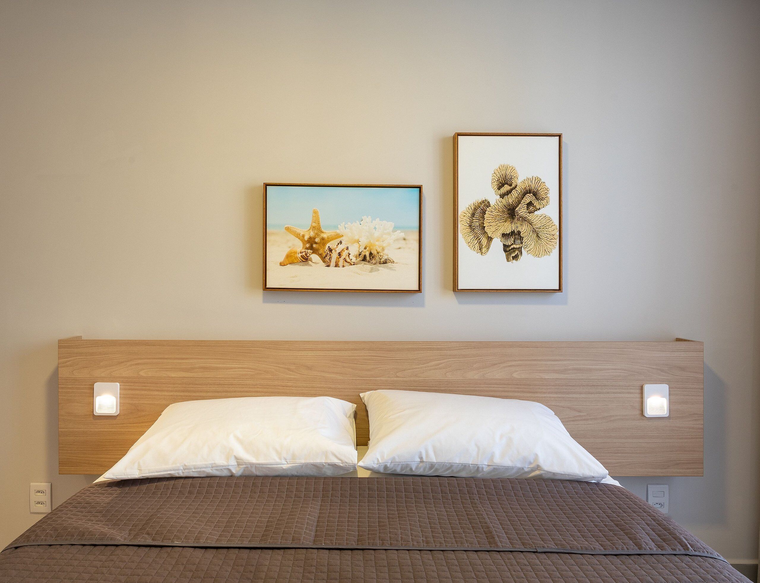 A bed with a quilted brown cover, two white pillows, and a wooden headboard. Two framed artworks are hung on the wall above the bed.