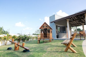 A playground with wooden structures, including a seesaw and a small house-like playhouse, in a grassy area with a building in the background.