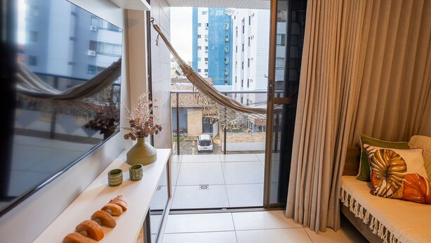 A modern living room with a large window, a sofa with cushions, a small table with decorative items, and a view of a courtyard with a hammock and a parked car. - Enseada de Coqueirinho #201 - Apt by Carpediem (João Pessoa)