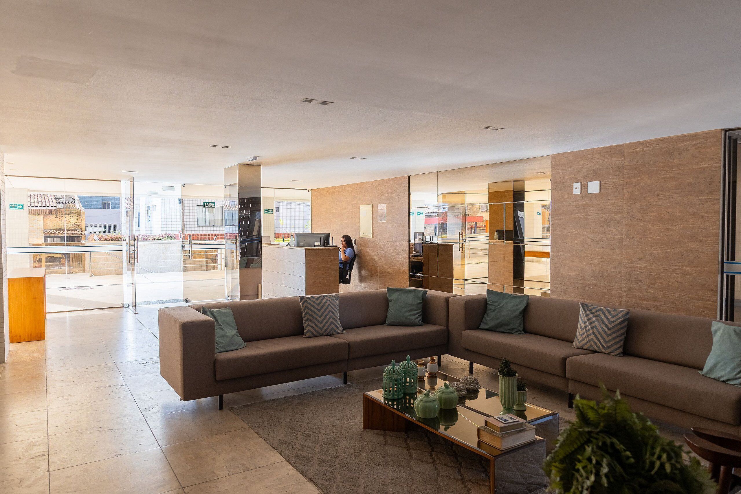Um lobby moderno com sofá em formato de L, mesa de centro com objetos decorativos e divisória de vidro.
