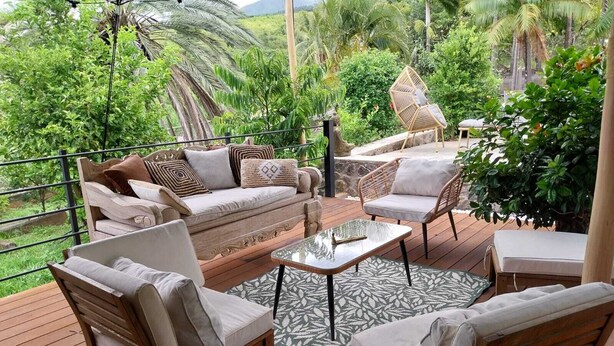 An outdoor deck with a sofa, chairs, and a table, surrounded by lush greenery and palm trees. - Modern Creole house with swimming pool, garden and sea view (Etang Salé les Hauts) (L'Étang-Salé)