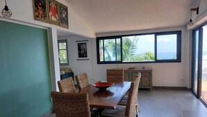 A dining area with a large table and wicker chairs, a cabinet with a green door, and a view of the outdoors through large windows. - Modern Creole house with swimming pool, garden and sea view (Etang Salé les Hauts) (L'Étang-Salé)