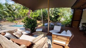 An outdoor lounge area with wooden furniture, a pool, and lush greenery in the background. - Modern Creole house with swimming pool, garden and sea view (Etang Salé les Hauts) (L'Étang-Salé)
