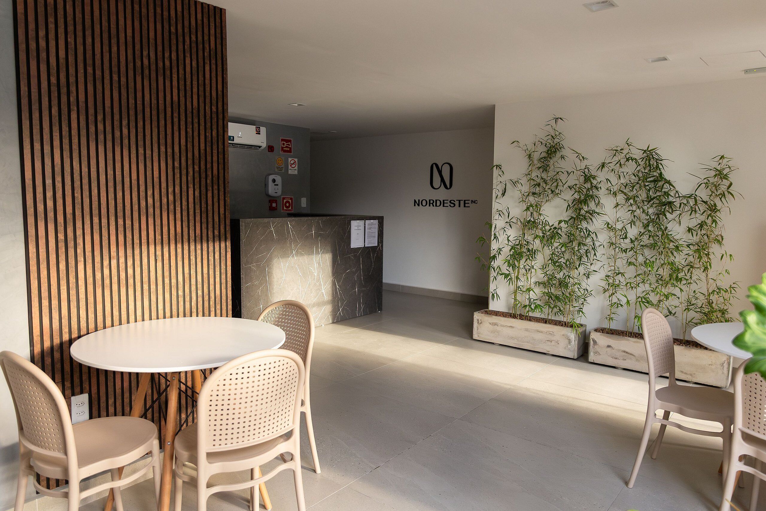 Uma área de recepção moderna, com balcão, mesa redonda com cadeiras e plantas de bambu ao fundo.