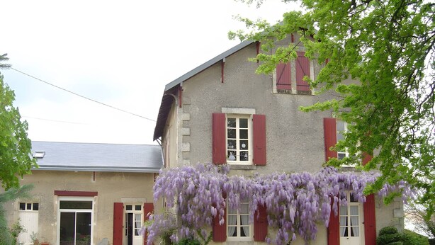 Un bâtiment à deux étages, doté d’une façade en pierre, de volets rouges et d’un porche couvert orné de glycines violettes.