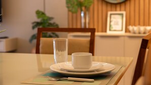 Ein Esstisch mit einem weißen Teller, einer weißen Tasse und einem Glas. Im Hintergrund steht ein Holzstuhl und an der Wand steht eine Pflanze.