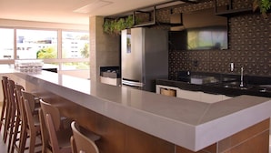 A modern kitchen with a large island, bar stools, a built-in oven, and a large window providing a view of the city. - Puerto Ventura #203 - Ape Ao Mar por Carpediem (João Pessoa)