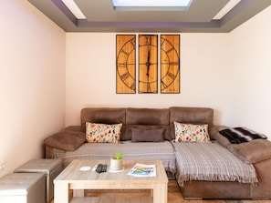 Ein Wohnzimmer mit einem braunen Sofa, zwei Zierkissen, einem Couchtisch mit Fernbedienungen und einer Zeitschrift sowie einer Wanduhr.