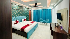 A modern hotel room with a bed, a desk, a chair, a ceiling fan, and a TV mounted on the wall. - The Prime Regency (Greater Noida)