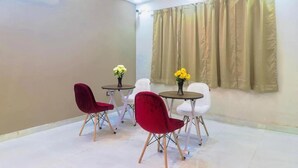 A room with a white tiled floor, a beige wall, and a large window with beige curtains. There are two round tables with chairs, one with red chairs and one with white chairs. - The Prime Regency (Greater Noida)