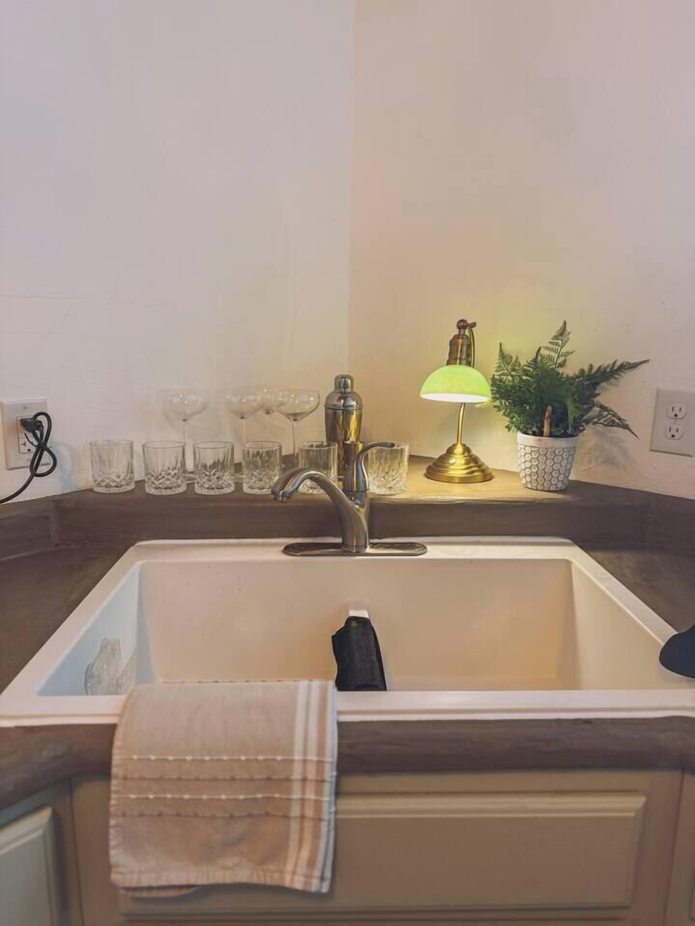 A kitchen sink with a towel hanging on the door, a green lamp, and a potted plant on the counter.