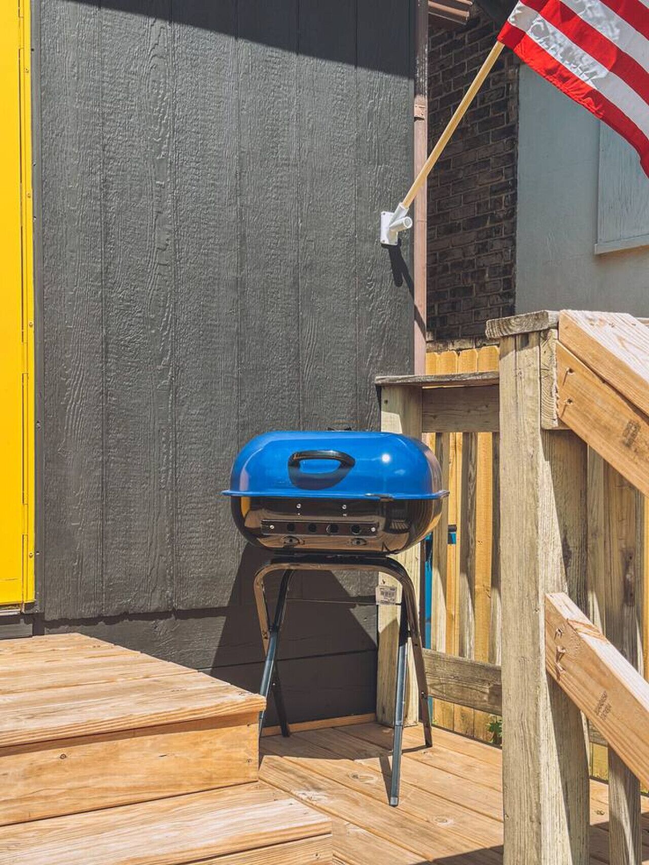 A blue barbecue grill on a wooden deck, a yellow door, and a wooden fence.