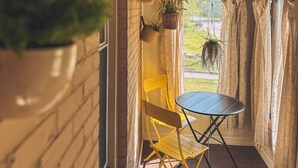 A small, enclosed porch with a brick wall, a yellow chair, a round table, and hanging planters. - The Row House on 4th (Dowagiac)