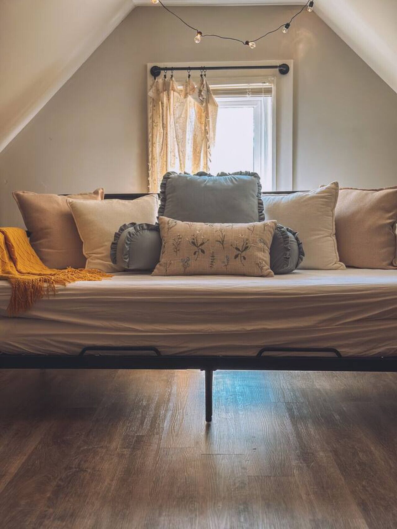 Een slaapkamer in een loft met een schuine plafond, een bed met veel kussens, een raam met gordijnen en lichtjes.