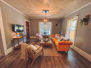 Un salon avec un plancher en bois, un canapé, un fauteuil, une table basse, un téléviseur et une fenêtre avec des rideaux.