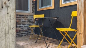 A wooden deck with two yellow chairs and a table, a brick wall, and large windows. - The Row House on 4th (Dowagiac)