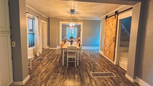 A spacious dining area with a wooden table and chairs, a chandelier, and a view through a window. - The Row House on 4th (Dowagiac)