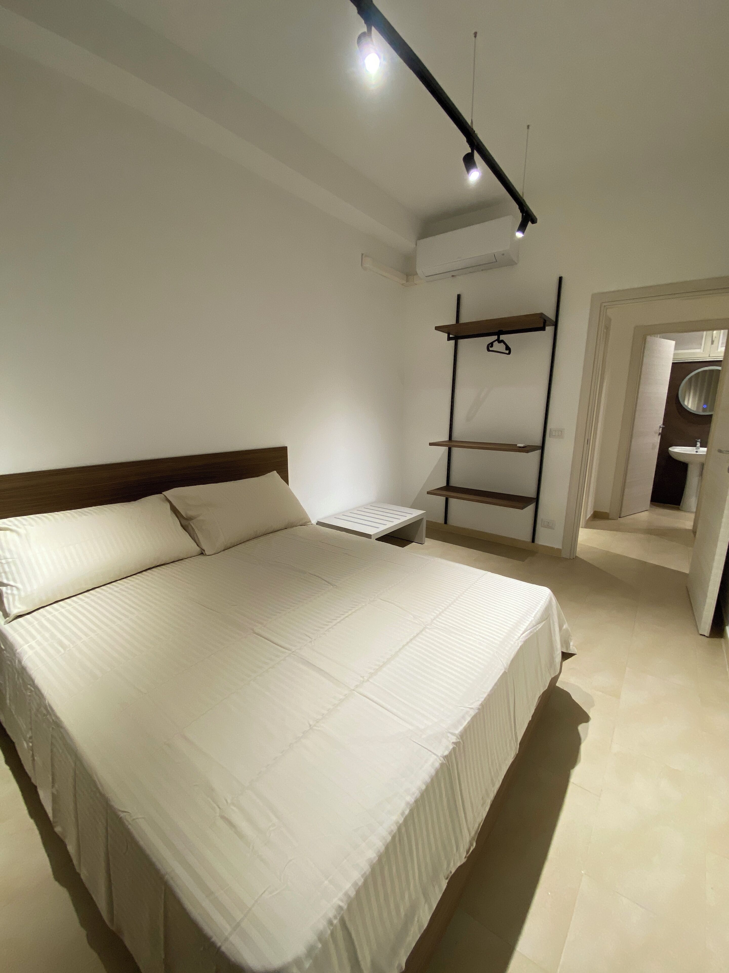 A bedroom with a bed, a bedside table, a hanging shelf, and an air conditioning unit.