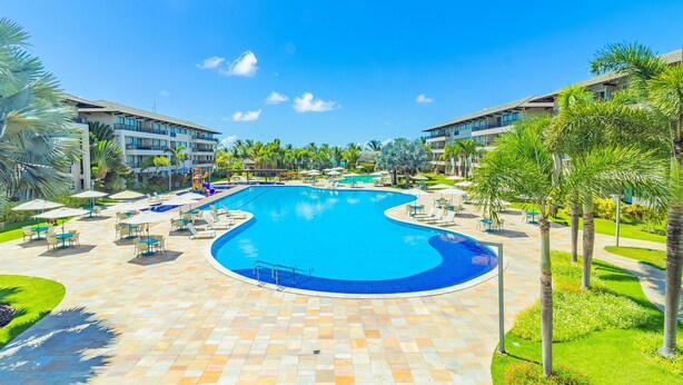 Uma grande piscina com design curvo, rodeada por espreguiçadeiras e guarda-sóis, com palmeiras e um céu azul límpido ao fundo.