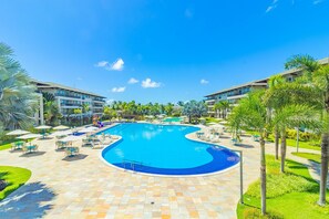A large swimming pool with a curved design, surrounded by lounge chairs and umbrellas, with palm trees and a clear blue sky in the background. - Eco Life #105 - Apt With Balcony by Carpediem (Ipojuca)