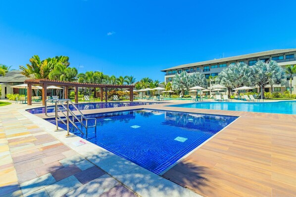 A swimming pool with a wooden deck and lounge chairs, surrounded by palm trees and a hotel building. - Eco Life #105 - Apt With Balcony by Carpediem (Ipojuca)