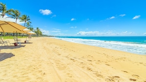 Uma praia com areal arenoso, palmeiras e cadeiras de praia sob um céu azul claro.