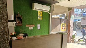 Un comptoir d’accueil d’hôtel avec un décor de fond en gazon artificiel vert, un panneau indiquant qu’il est interdit d’apporter de la nourriture de l’extérieur, et un petit autel orné de fleurs.