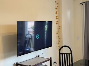 Un televisor de pantalla plana montado en la pared, una mesa de comedor con sillas y una cortina a la derecha.