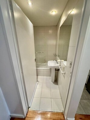 Een smalle badkamer met een witte wastafel, een spiegel en een douchehoek.