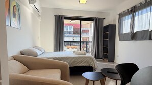 Ein modernes Schlafzimmer mit Bett, einer Couch, einem kleinen Tisch und einem Stuhl. Ein Fenster mit Vorhängen, eine Deckenleuchte und eine Wand-Klimaanlage sind vorhanden.