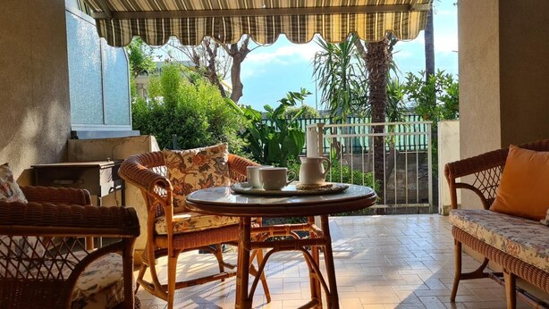Uma varanda coberta com móveis de vime, uma mesa redonda e vista para vegetação e um edifício ao fundo.