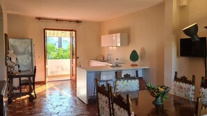 Uma cozinha moderna com ilha central, área de jantar com cadeiras florais e vista para o verde pela porta.