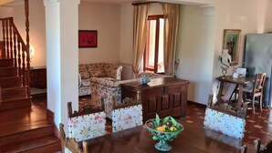 Uma sala de estar ampla com escada de madeira, um conjunto de sofás, uma mesa de jantar com um vaso de flores e um quadro na parede.