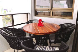 A round wooden table with four black wicker chairs, a red decorative item, and a window with a view of buildings. - Mont Cristo #401 - Beachfront por Carpediem (João Pessoa)
