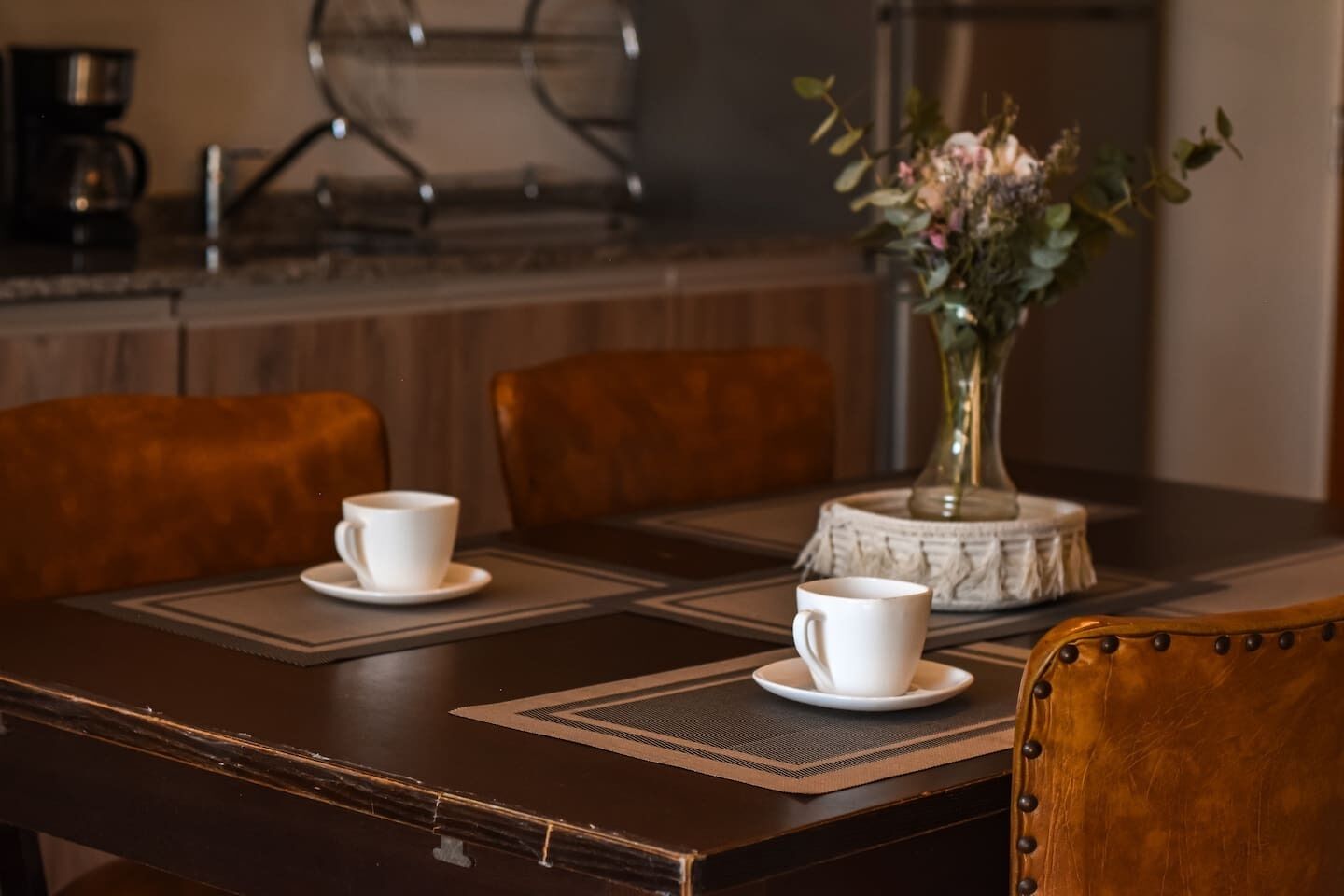 A dining table with two white cups and saucers, a vase with flowers, and a coffee machine in the background.