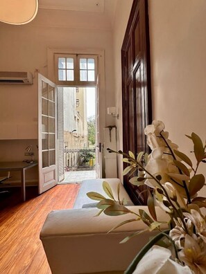 Um quarto com piso de madeira, um banco branco, uma planta em vaso e vista através de uma porta aberta para uma varanda com guarda-corpo.