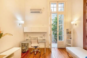 Quarto compacto e bem iluminado, com piso de madeira, uma mesa com cadeira, uma estante e uma porta que dá para uma área externa.