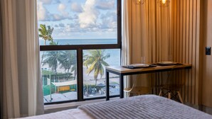 Een hotelkamer met een groot raam met uitzicht op een strand, een houten bureau en een bed.
