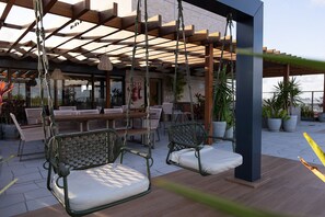 Ein überdachter Außenbereich mit Korbstühlen, einem Holztisch und üppigen Pflanzkübeln, umgeben von einer Pergola.