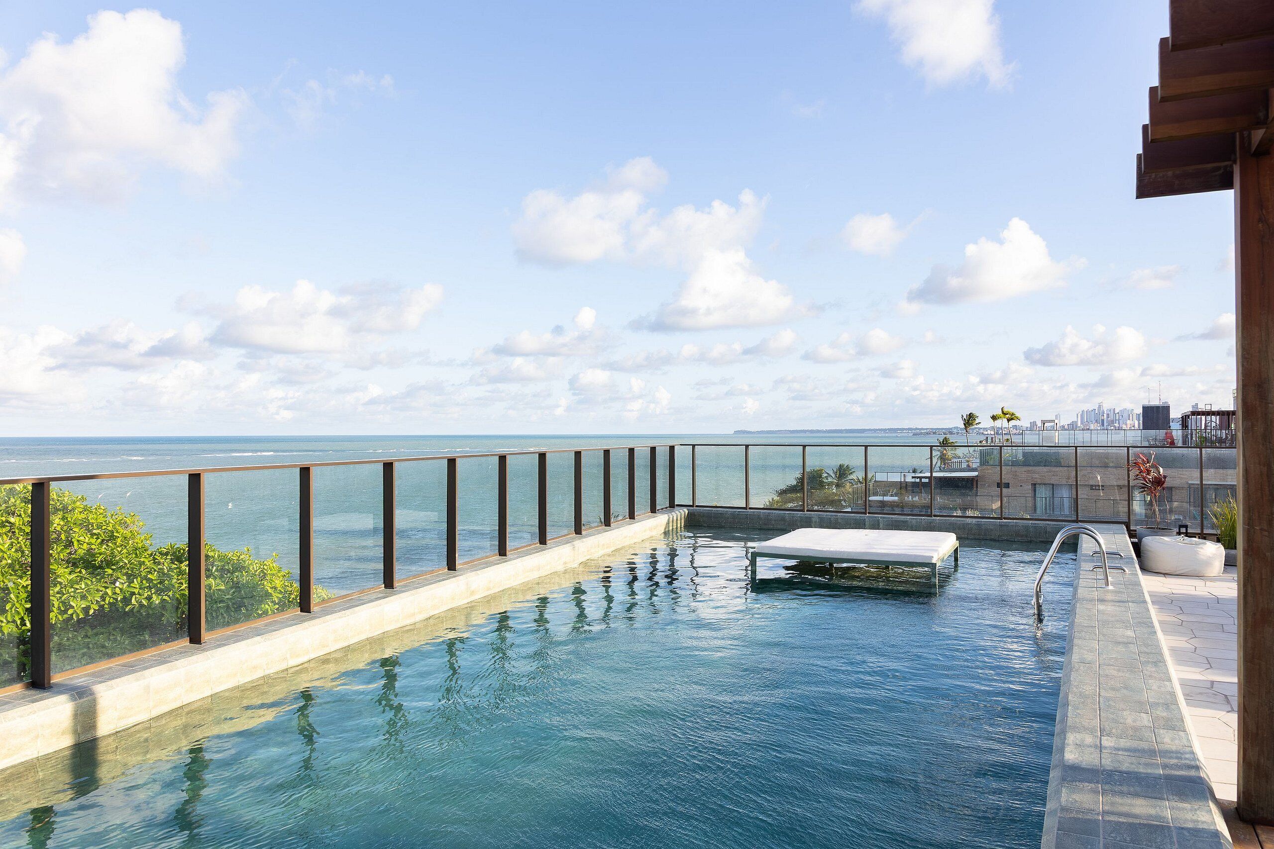 A rooftop infinity pool with a glass railing, a small white lounge chair, and a view of the ocean and city skyline.