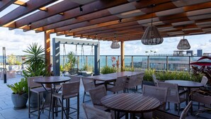 Een terras buiten met houten tafels en rieten stoelen, potplanten en hangende rieten lampen.
