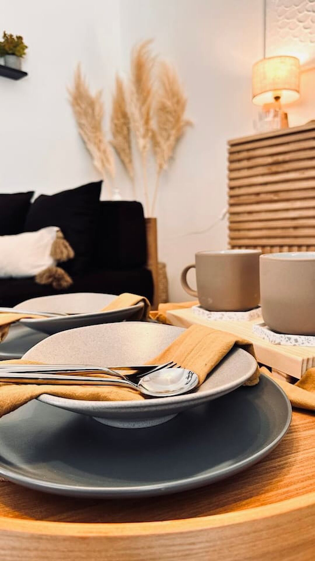 A dining table with plates, cutlery, and a cup, set against a wall with pampas grass and a lamp.