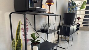 A modern, black metal shelf unit with multiple levels, holding various items like a potted plant, glassware, and a small table lamp. - Plaza Serrano Front Row Loft by airesB&B (Buenos Aires)