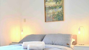 Ein ordentlich bezogenes Bett mit grauer Bettdecke und Kissen, ein weißer Nachttisch mit einer Lampe sowie ein Gemälde an der Wand.