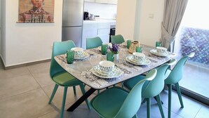 A dining area with a table set for four, green chairs, a clock on the wall, and a kitchen in the background. - Luxury 2BR Apt at Elysia Park S6 with Pools, Gym & Spa - Center Paphos (Pafos)